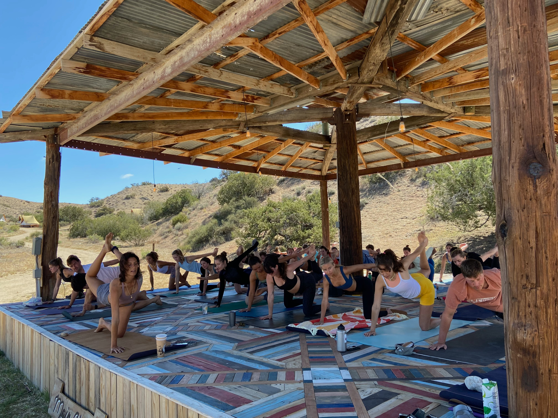 Group yoga class under an open-air wooden pavilion in the hills